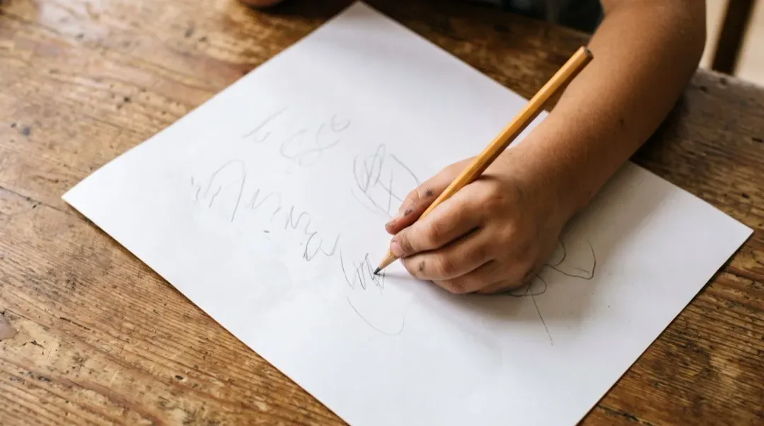 Ein schräg auf einem Tisch liegendes Blatt Papier, auf das eine linke Kinderhand eine Zahl schreibt.